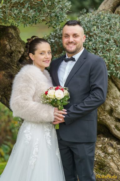 Un couple heureux en tenue de mariage, posant près d'un arbre, avec un bouquet de fleurs.
