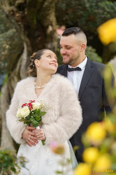 Un couple en tenue de mariage, souriant, au milieu de la nature, avec des fleurs à la main.